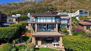 Une maison moderne à deux étages, dotée d’un balcon, entourée d’une végétation luxuriante et avec une chaîne de montagnes en toile de fond.