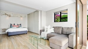 Une chambre moderne avec un lit, un canapé gris, une table basse en verre et une fenêtre donnant sur un paysage verdoyant.