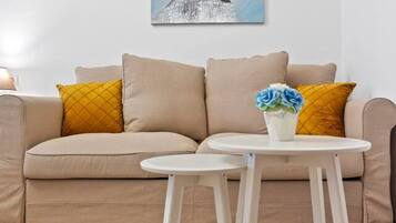 Una sala de estar con un sofá color beige, dos almohadas mostaza, una mesa de centro blanca con un jarrón de flores azul, y un cuadro en la pared.