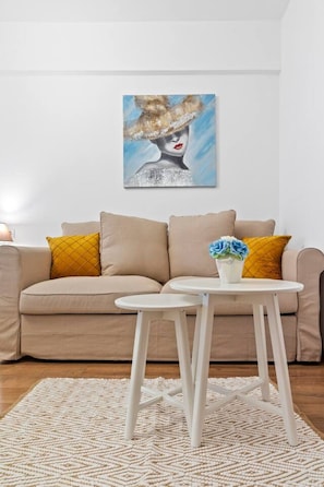 A living room with a beige sofa, two mustard pillows, a white coffee table with a blue flower vase, and a painting on the wall.