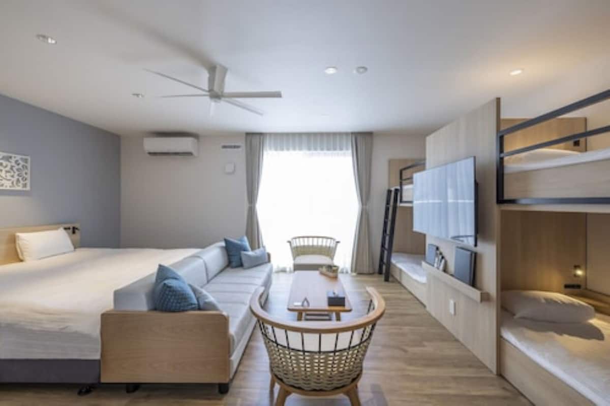 A modern hotel room with a bed, sofa, dining table, and a TV mounted on the wall.