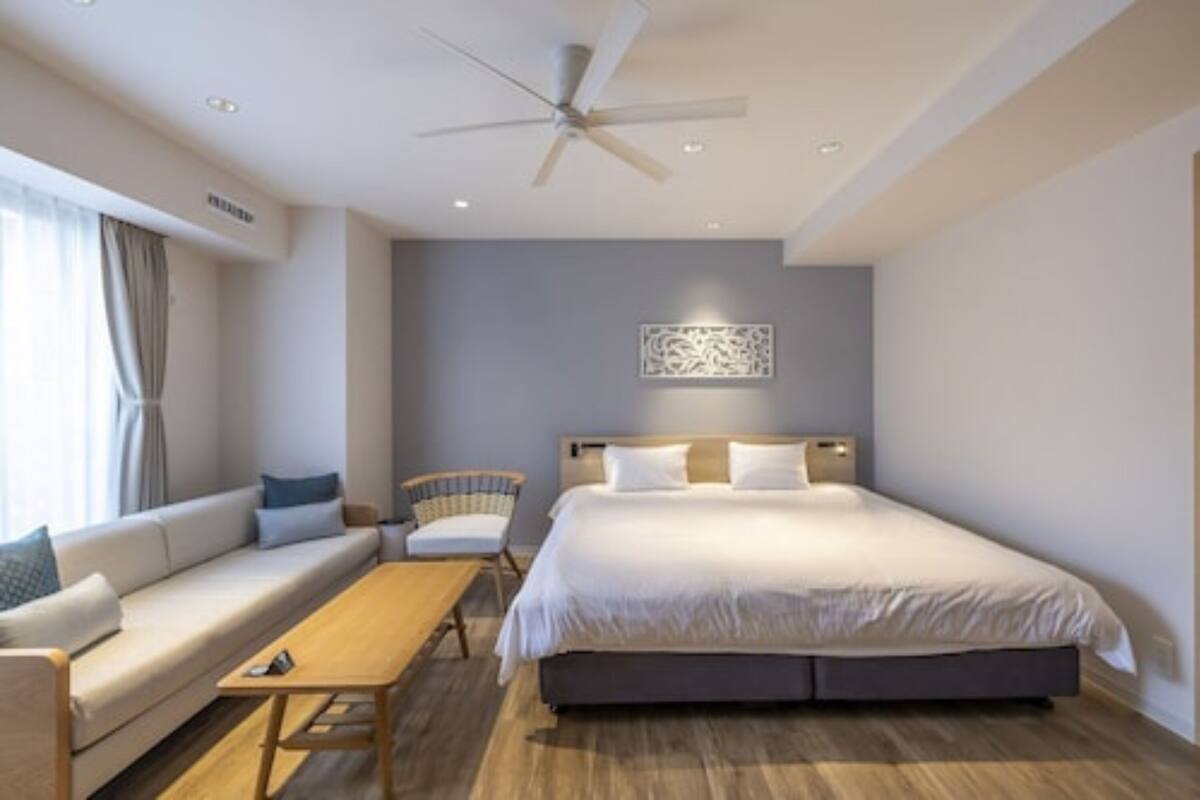 A modern hotel room with a large bed, a sofa, a wooden table, and a ceiling fan.
