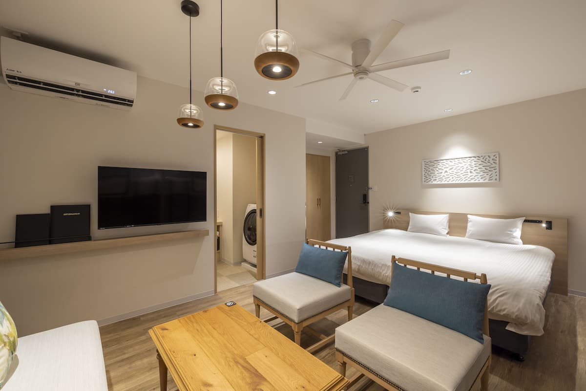 A modern hotel room with a bed, two chairs, a TV, and a ceiling fan.