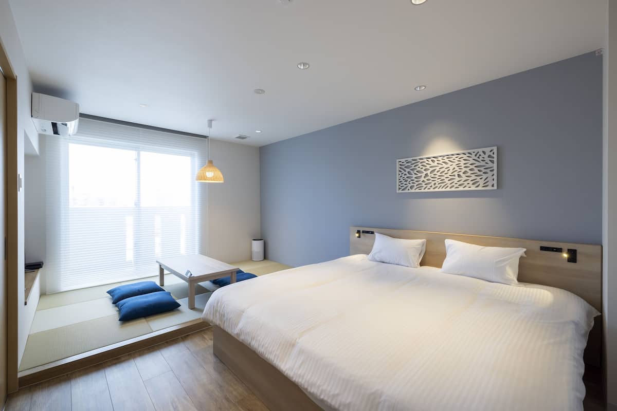 A modern hotel room with a large bed, a seating area with cushions, a window with blinds, and a decorative wall piece.