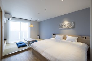 A modern hotel room with a large bed, a seating area with cushions, a window with blinds, and a decorative wall piece.
