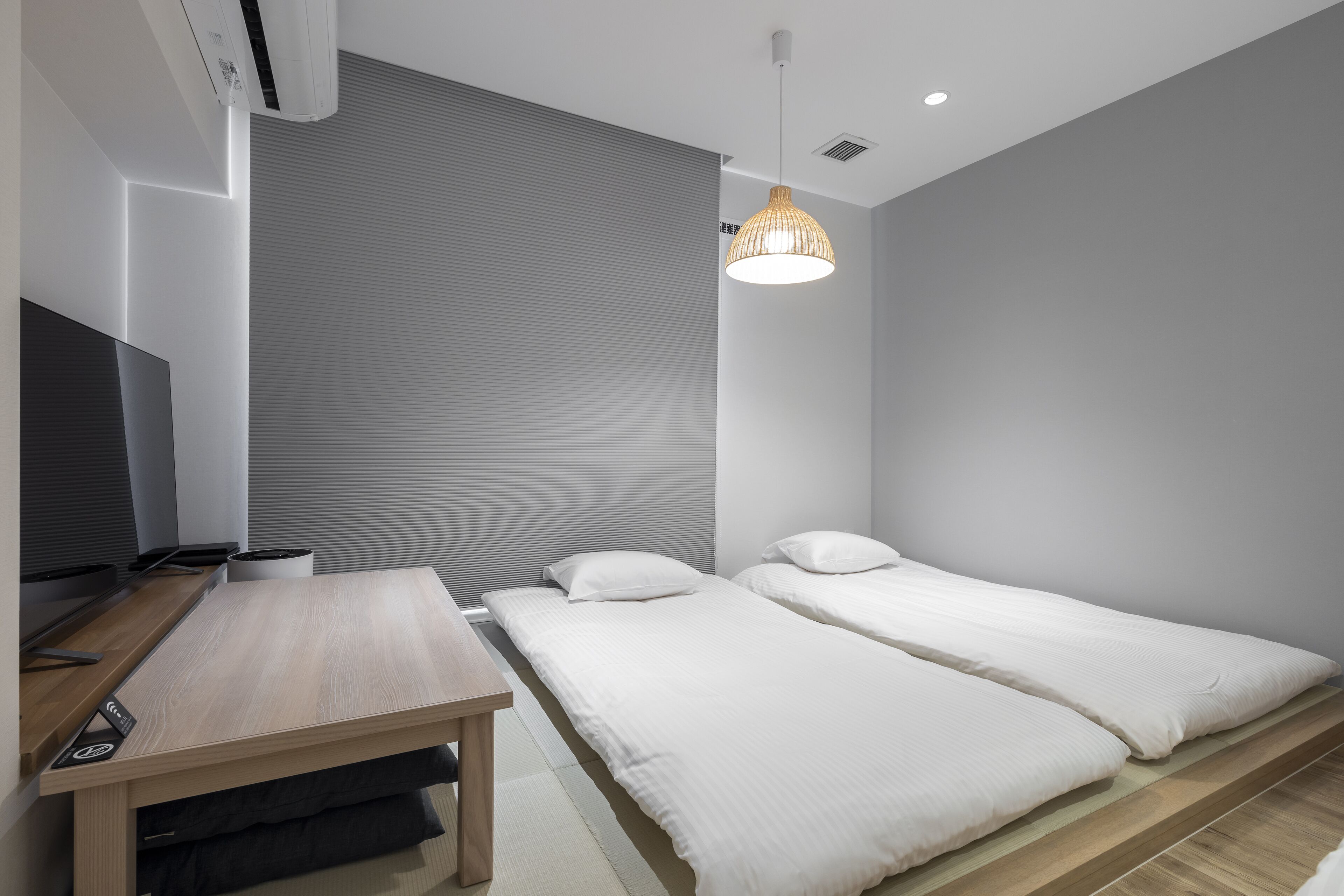 Quarto de hotel moderno com escrivaninha de madeira, duas camas de solteiro, uma janela grande com persianas e um lustre.