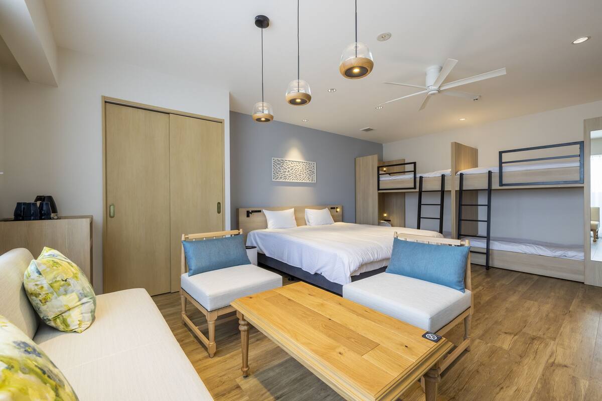A modern hotel room with a large bed, a sofa, a wooden table, and bunk beds in the background.