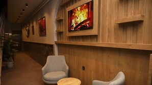 A hallway with wooden paneling, two grey armchairs, a small round table, and a wall-mounted TV displaying a fire. - Rush Otel Tokat (Tokat)