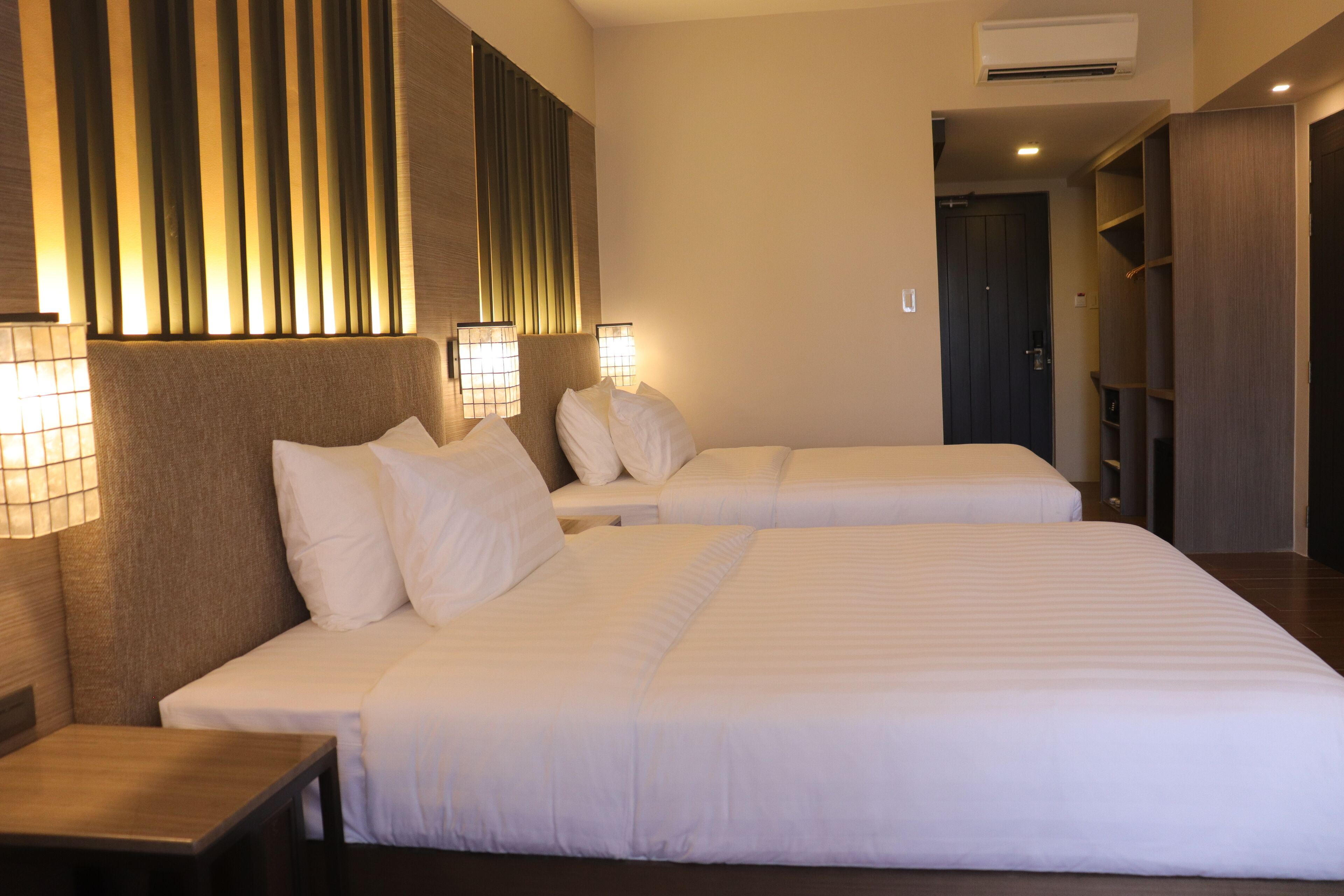 A hotel room with two beds, a nightstand, a wall-mounted lamp, and a wardrobe.