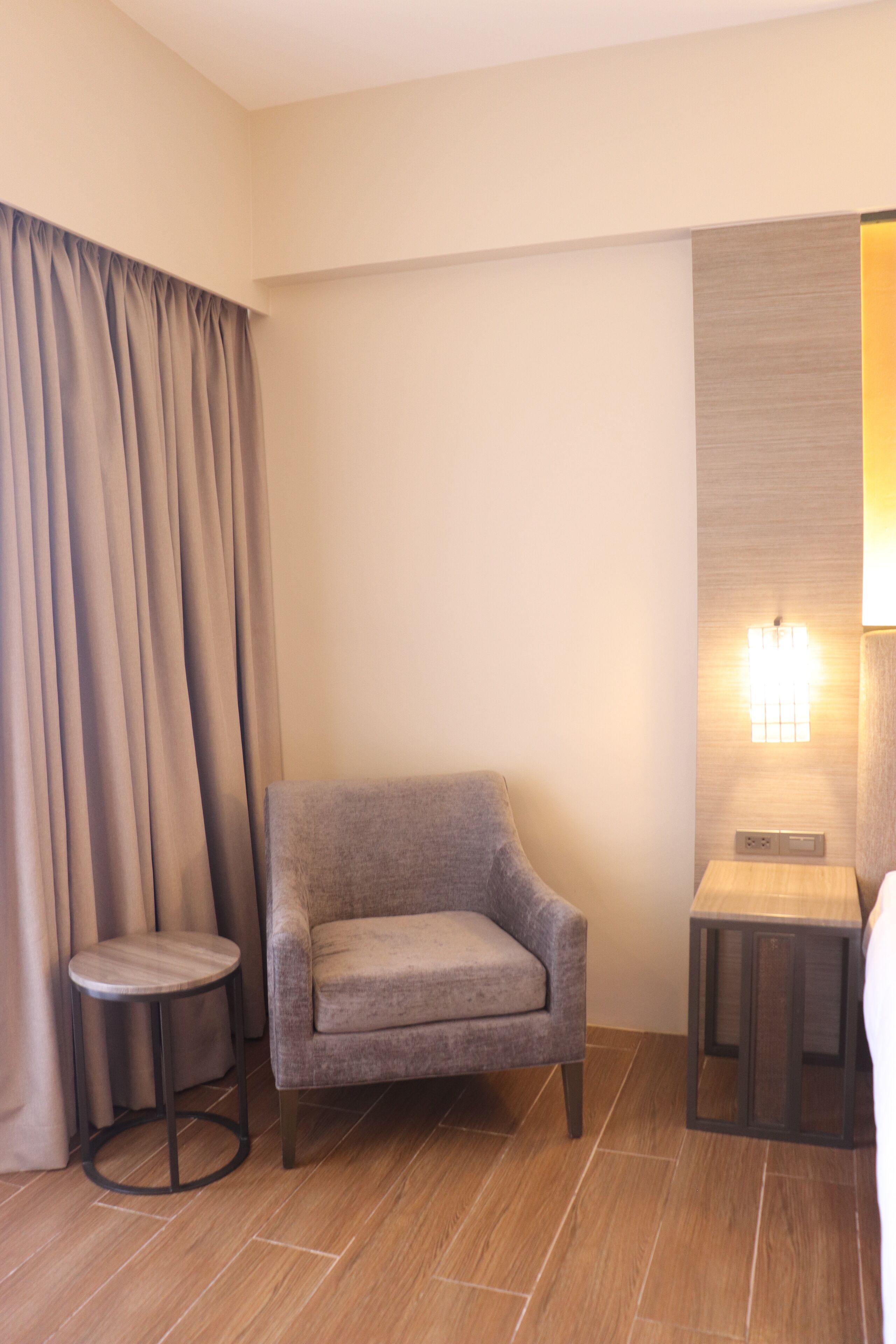 Una habitación de hotel con un sillón gris, una mesita redonda pequeña, una lámpara y una cortina.