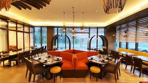 A restaurant with a central orange seating area, multiple dining tables, and large windows providing views. - The Aurora Subic Hotel Managed by HII (Olongapo)