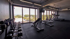 A gym with exercise equipment, large windows, and a view of a cityscape. - The Aurora Subic Hotel Managed by HII (Olongapo)