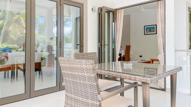 A modern dining area with a glass table and wicker chairs, a sliding glass door leading to a balcony, and a painting on the wall. - Palm Holiday Apartments (Praslin Island)