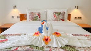 A neatly made bed with white linens, two pillows with tropical leaf patterns, and a towel arranged in the shape of swans with hibiscus flowers. - Palm Holiday Apartments (Praslin Island)