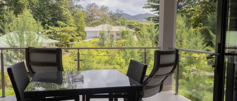 A balcony with a glass table and chairs, offering a view of lush greenery and distant buildings.