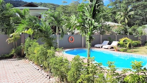 A swimming pool surrounded by lush greenery, a lifebuoy on a wall, and a tiled patio area. - Palm Holiday Apartments (Praslin Island)