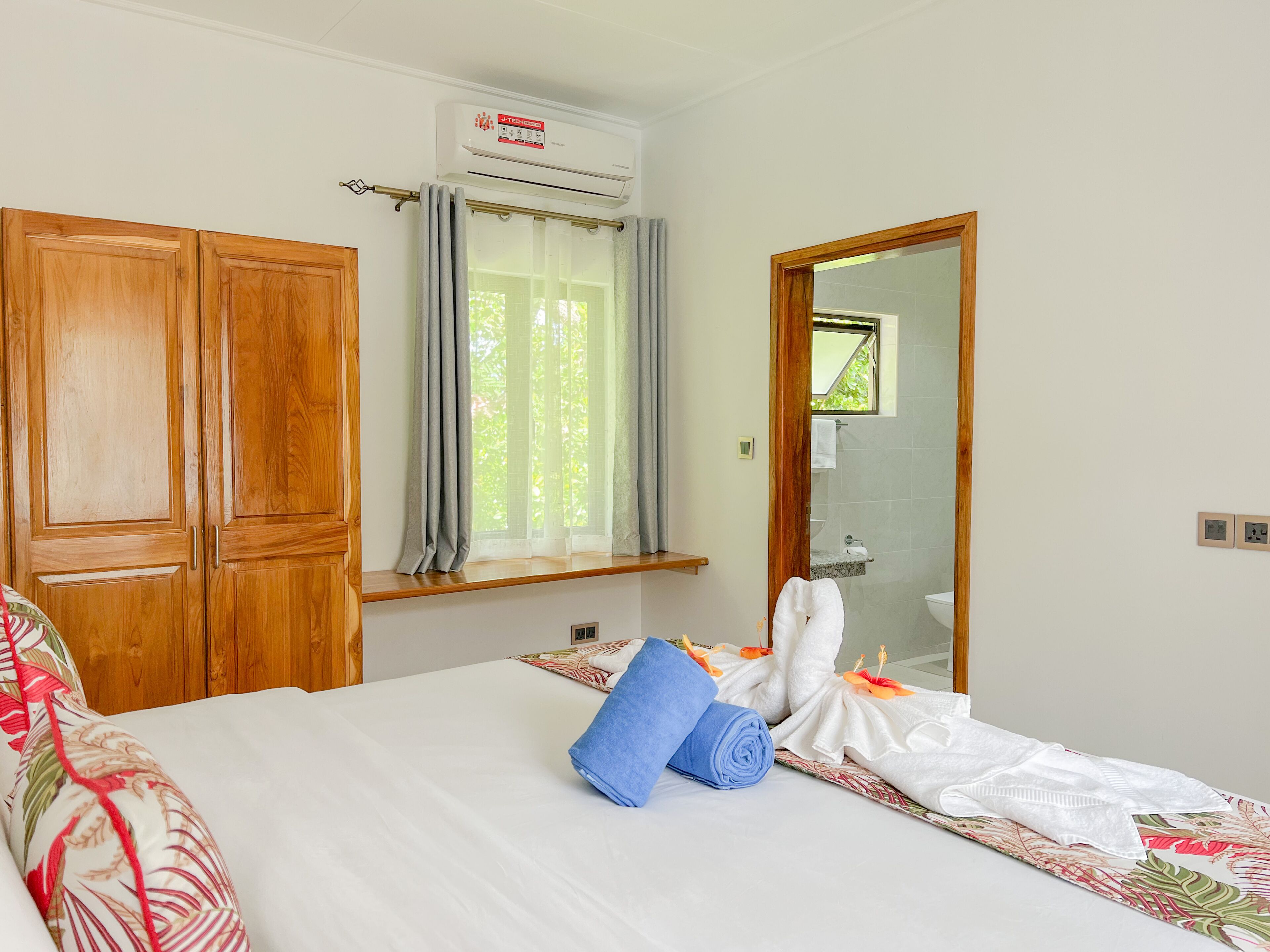 A hotel room with a bed, a wooden wardrobe, a window with curtains, an air conditioning unit, and a bathroom visible through a mirror.