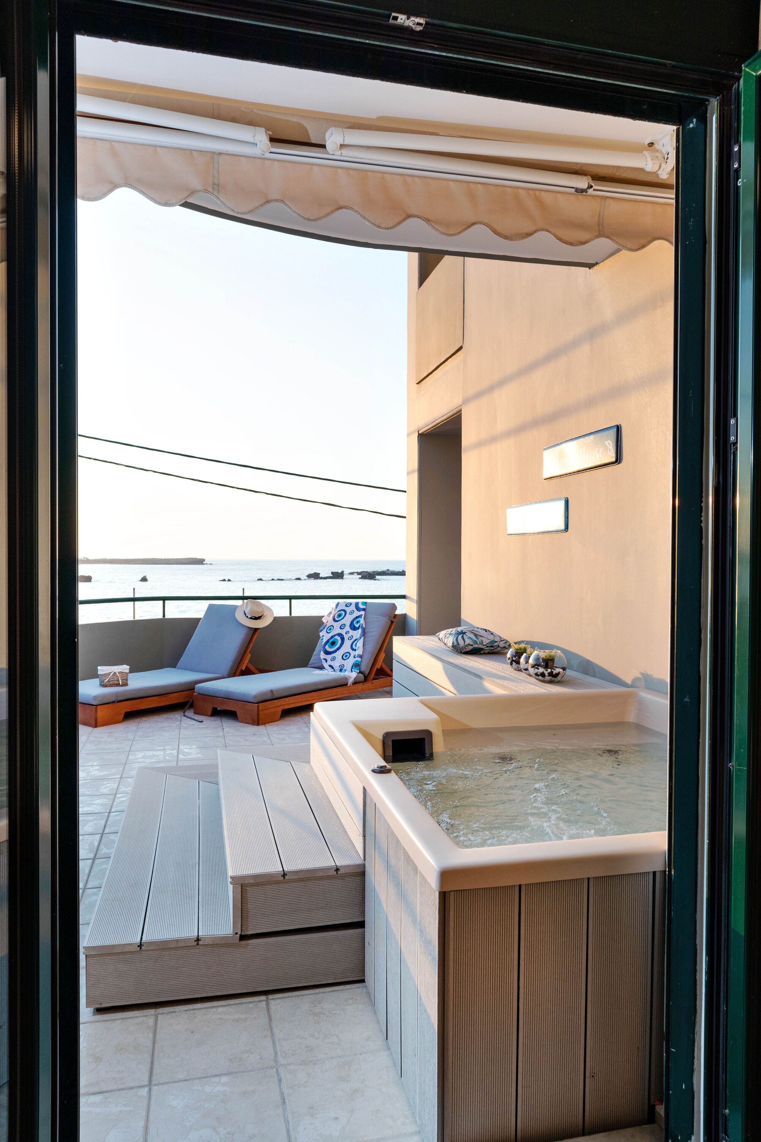 A balcony with two lounge chairs, a hot tub, and a view of the sea.