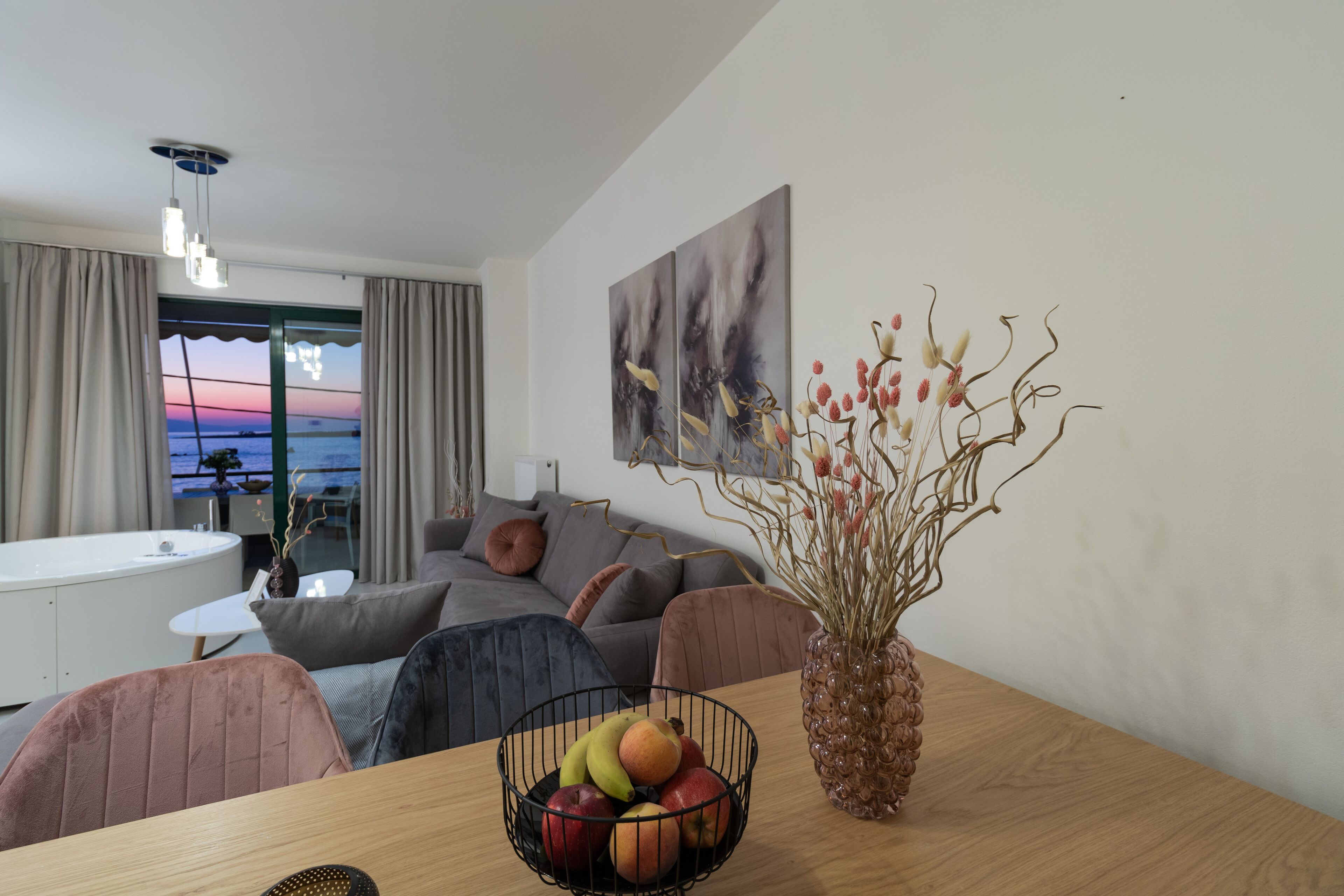 Een moderne woonkamer met een houten eettafel, een schaal met fruit, een vaas met gedroogde takken, een bank en uitzicht op zee door een groot raam.