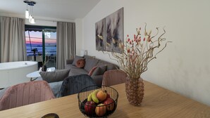 Een moderne woonkamer met een houten eettafel, een schaal met fruit, een vaas met gedroogde takken, een bank en uitzicht op zee door een groot raam.