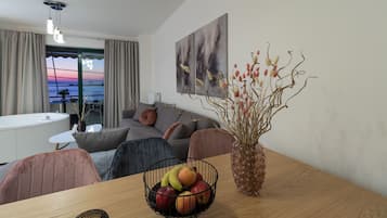 Een moderne woonkamer met een houten eettafel, een schaal met fruit, een vaas met gedroogde takken, een bank en uitzicht op zee door een groot raam.