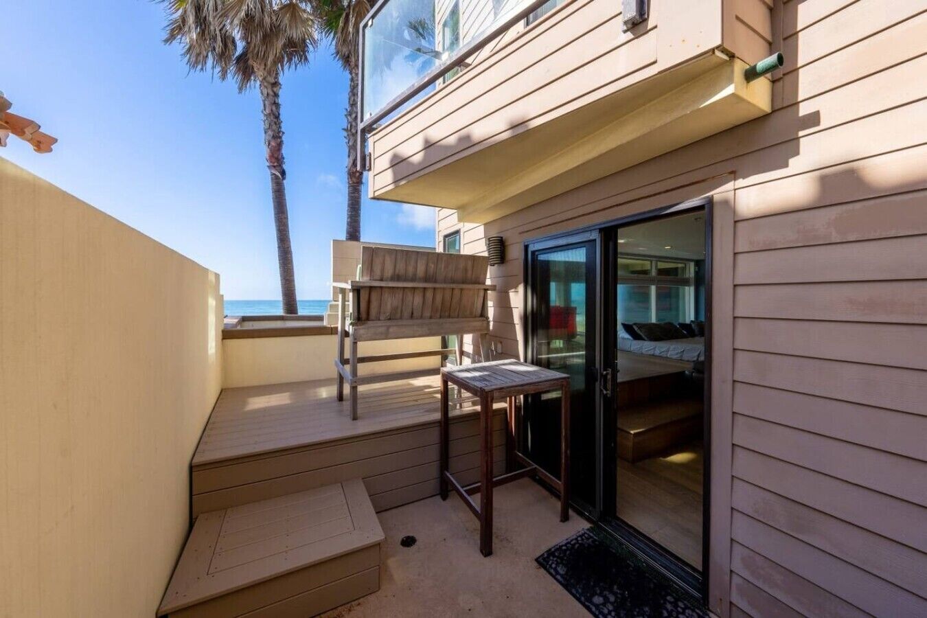 A balcony with a wooden bench, a small table, and a glass door leading to a bedroom.
