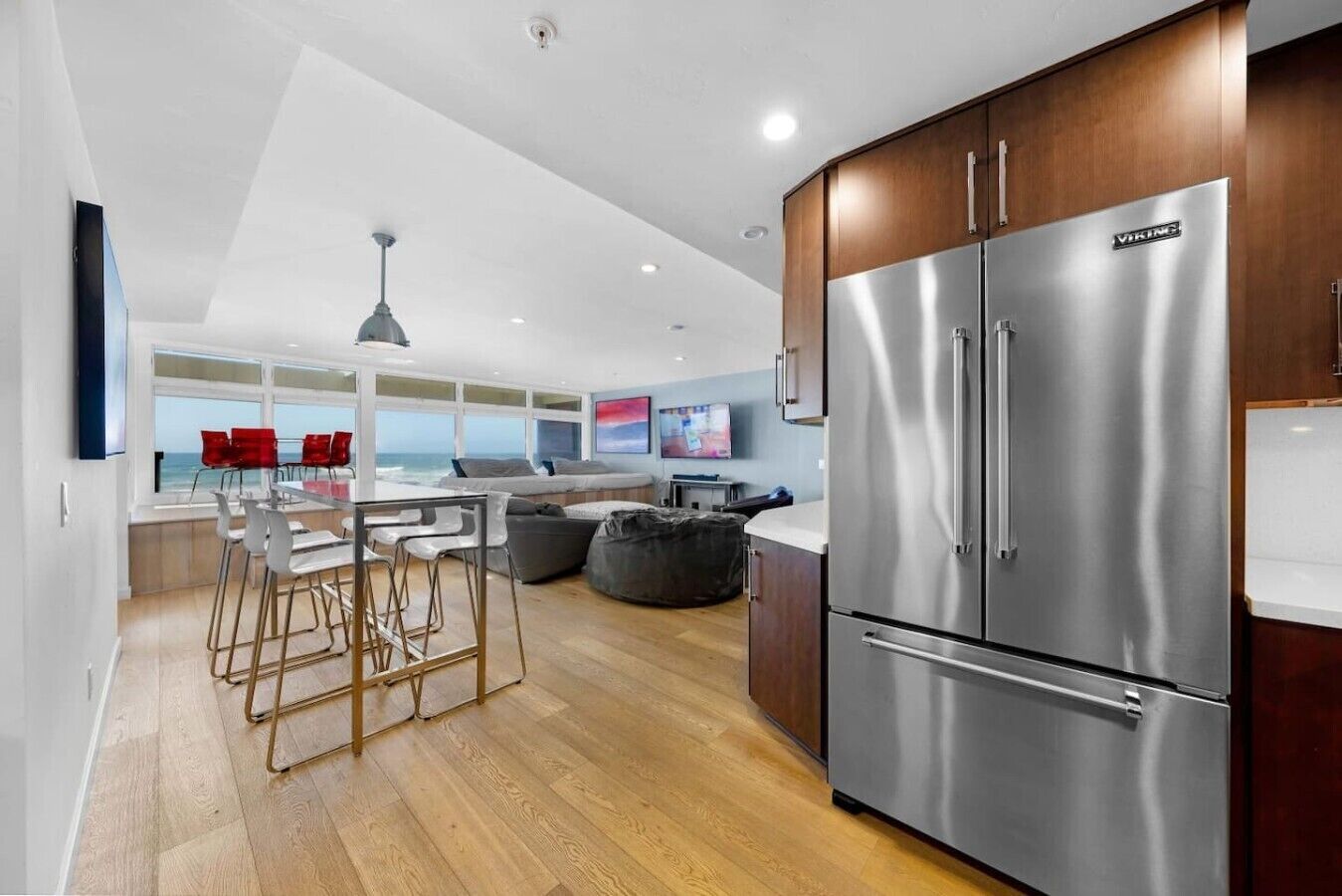A modern kitchen with a stainless steel refrigerator, wooden cabinets, and a dining area with bar stools.