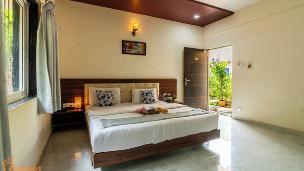 A bedroom with a bed, a nightstand, a wall-mounted lamp, a painting, and a door leading to a balcony with plants. - Arnnas Resort Diveagar (Shrivardhan)