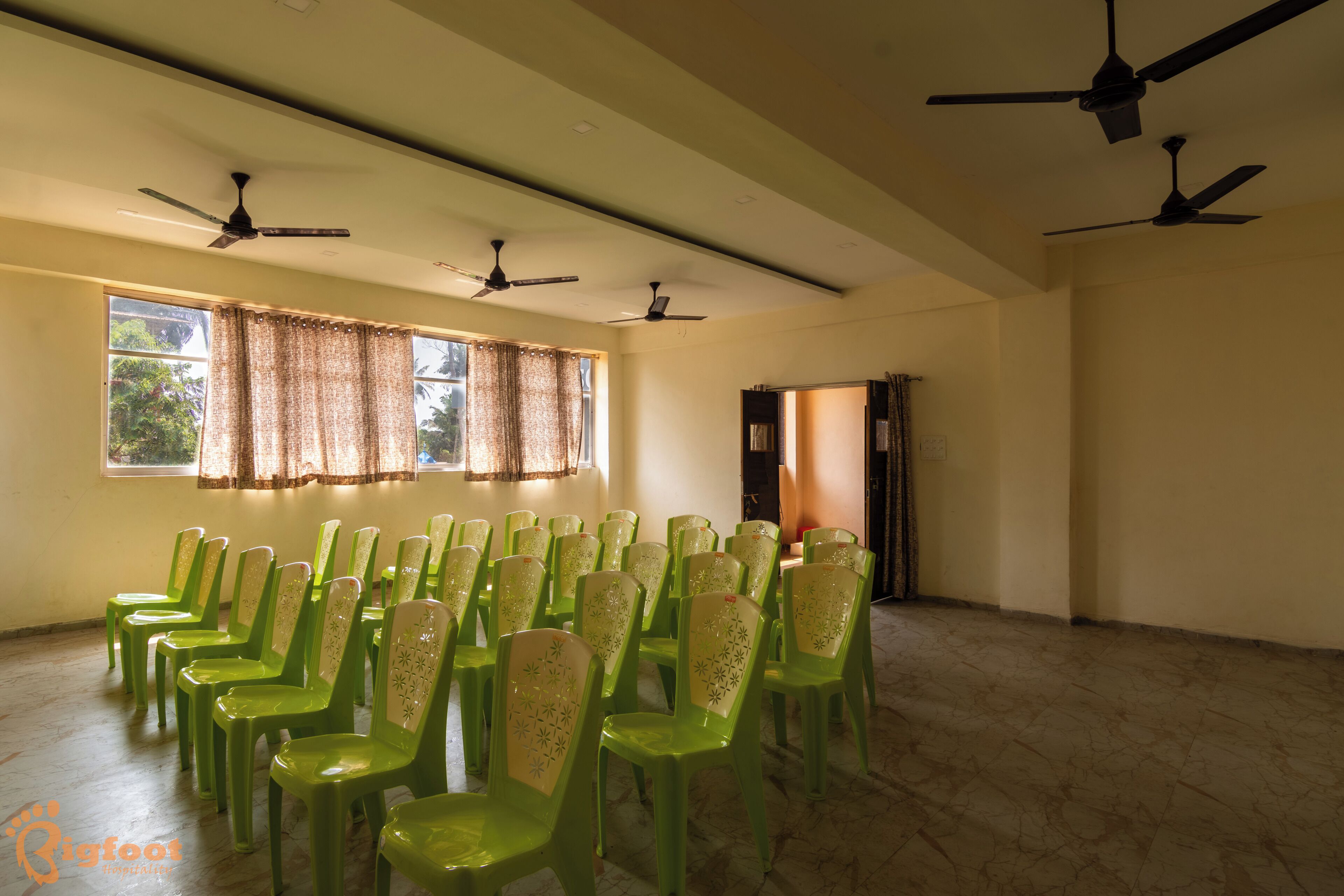 A room with multiple ceiling fans, green plastic chairs arranged in rows, and a window with curtains.