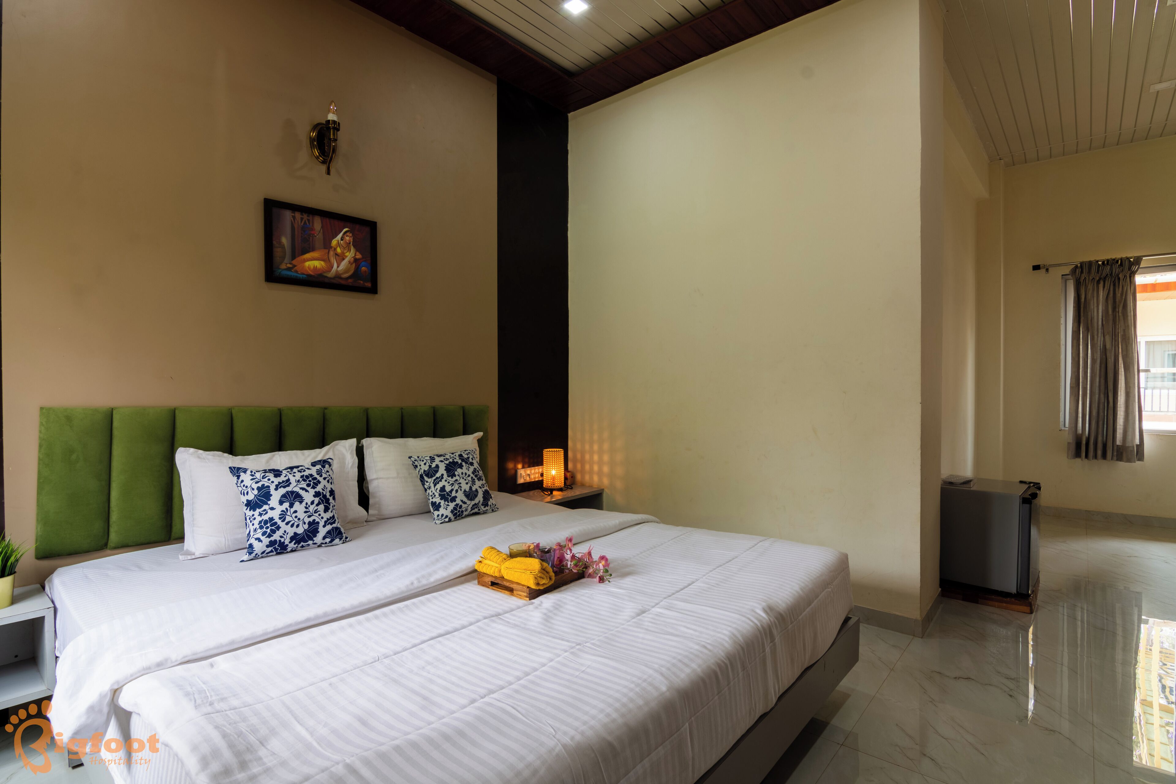 Habitación de hotel con una cama grande, una mesita de noche con lámpara, un cuadro en la pared y una ventana con cortinas.