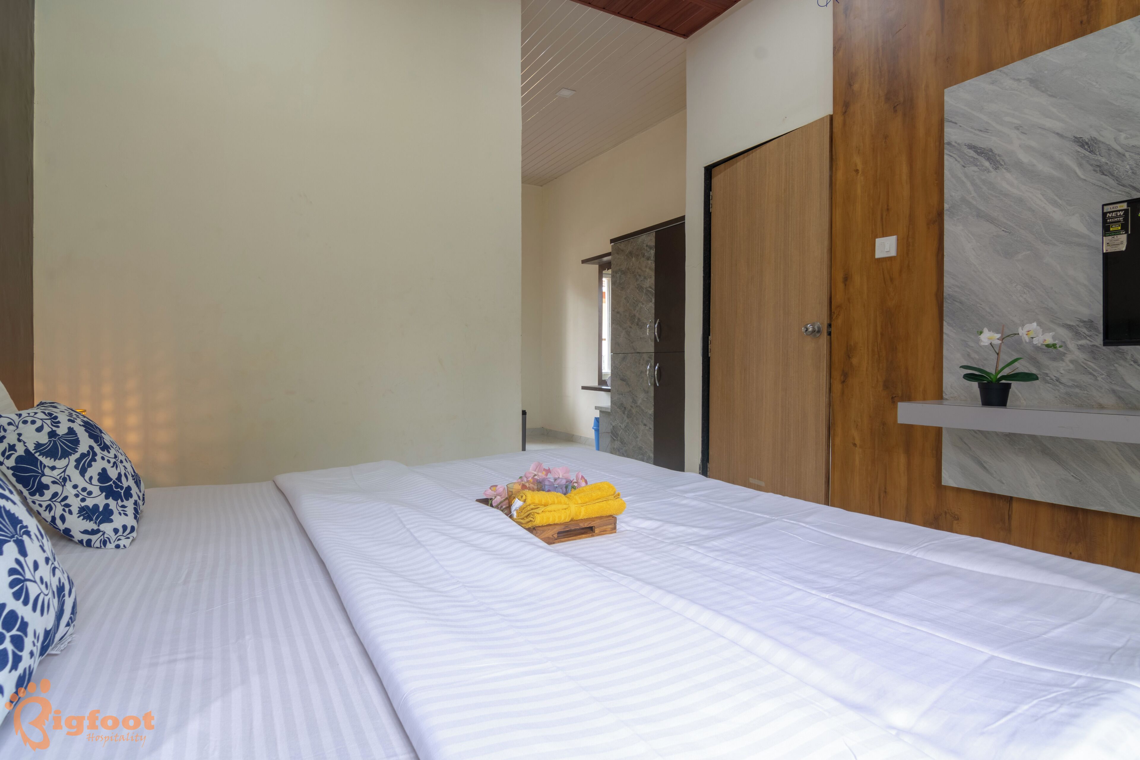 A neatly made bed with a wooden headboard, a small bedside table with a plant, and a marble wall mirror.