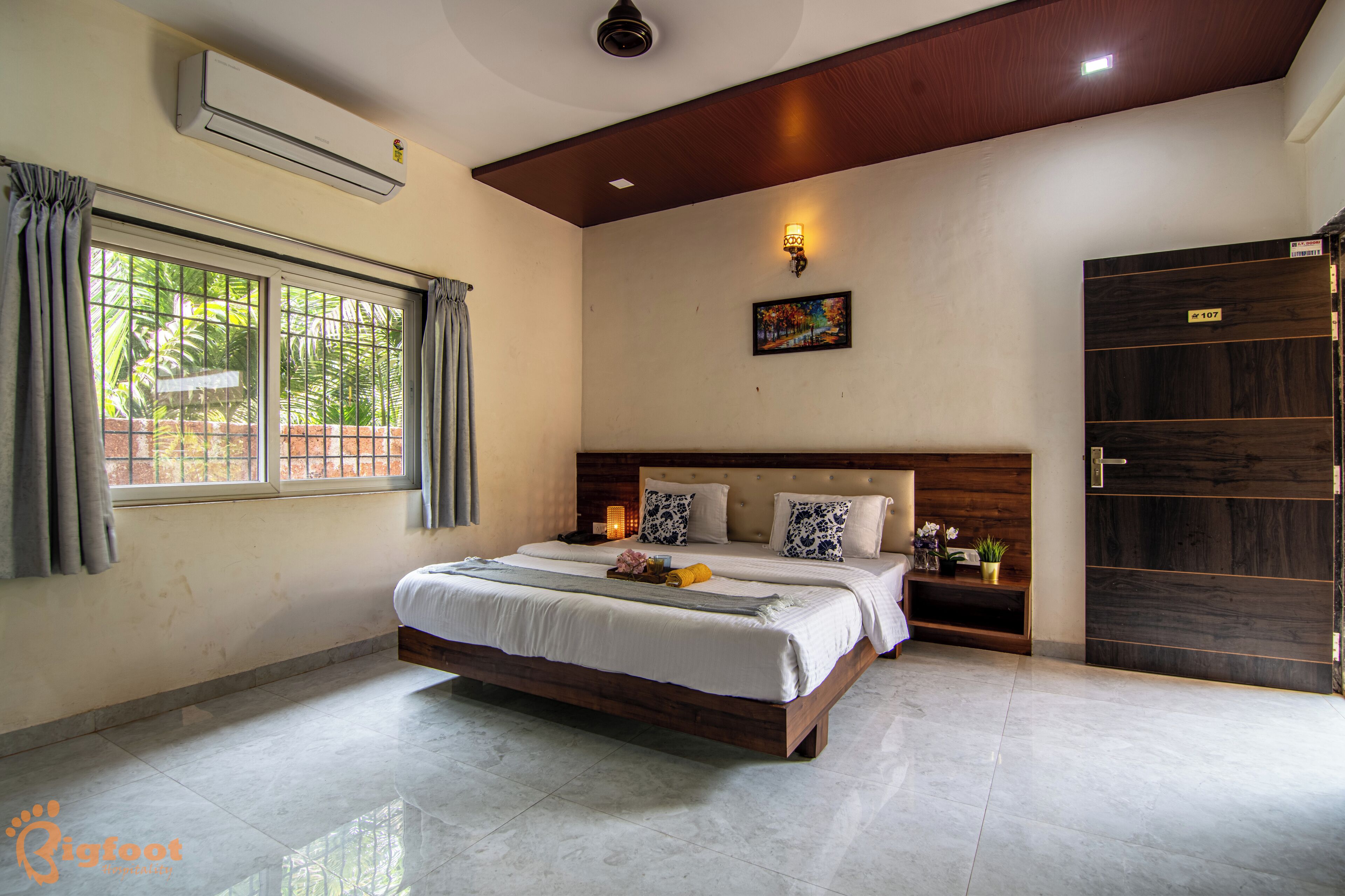 A bedroom with a large bed, a nightstand, a wall-mounted air conditioner, a window with curtains, and a door with a sign.