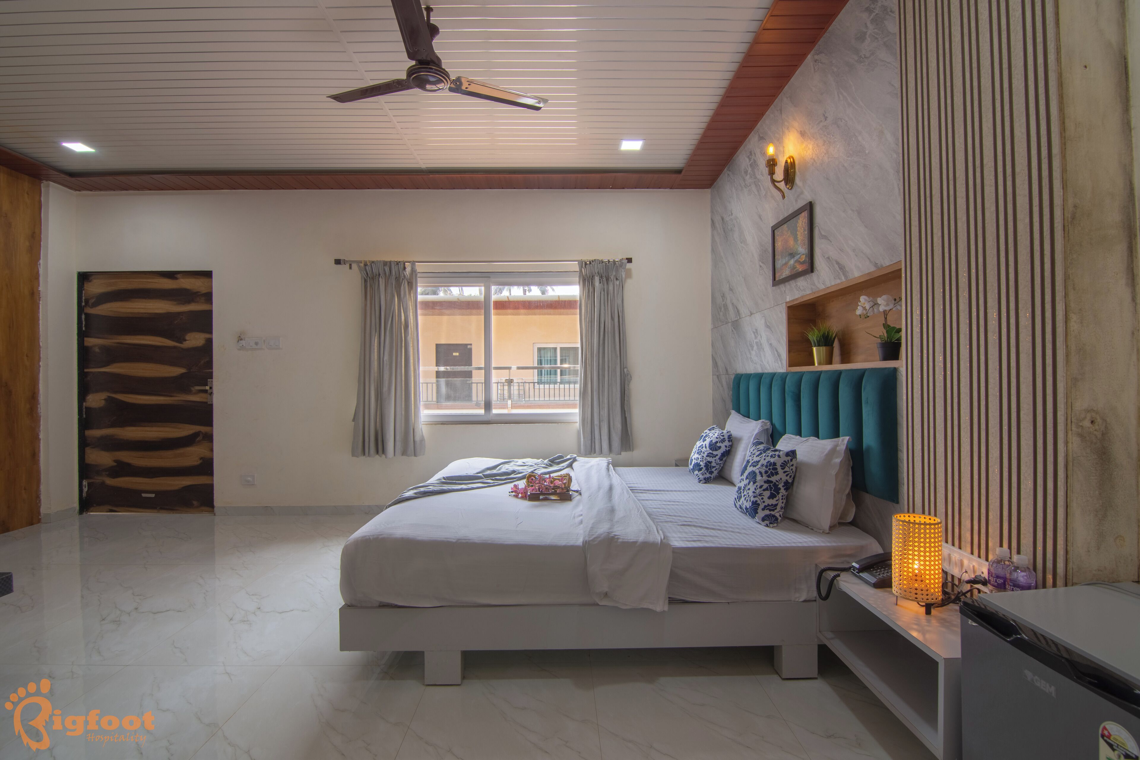 A bedroom with a bed, a ceiling fan, a window with curtains, a bedside table with a lamp, and a wall-mounted shelf with plants.