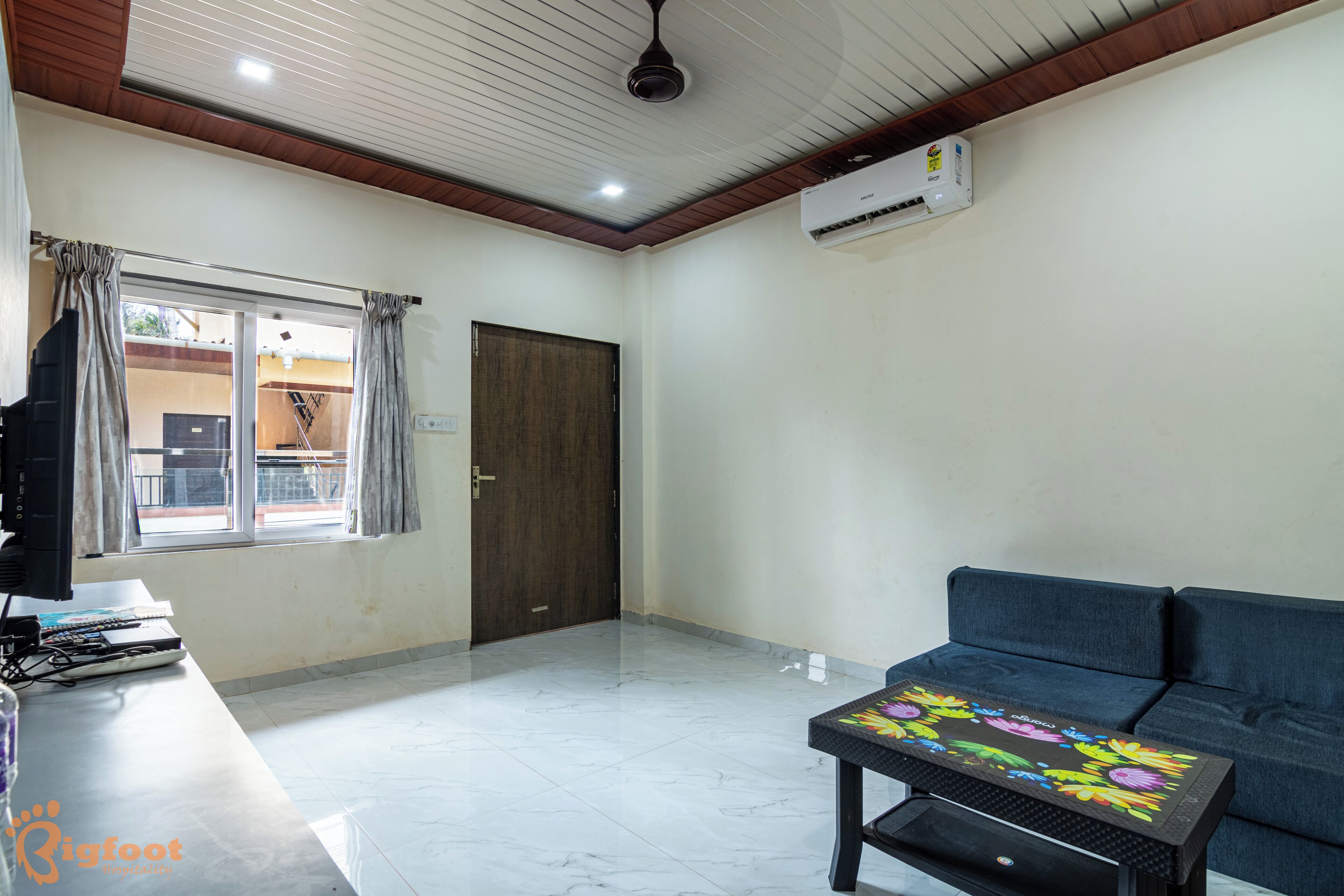 A room with a sofa, a table with a floral tablecloth, a TV, and an air conditioning unit.