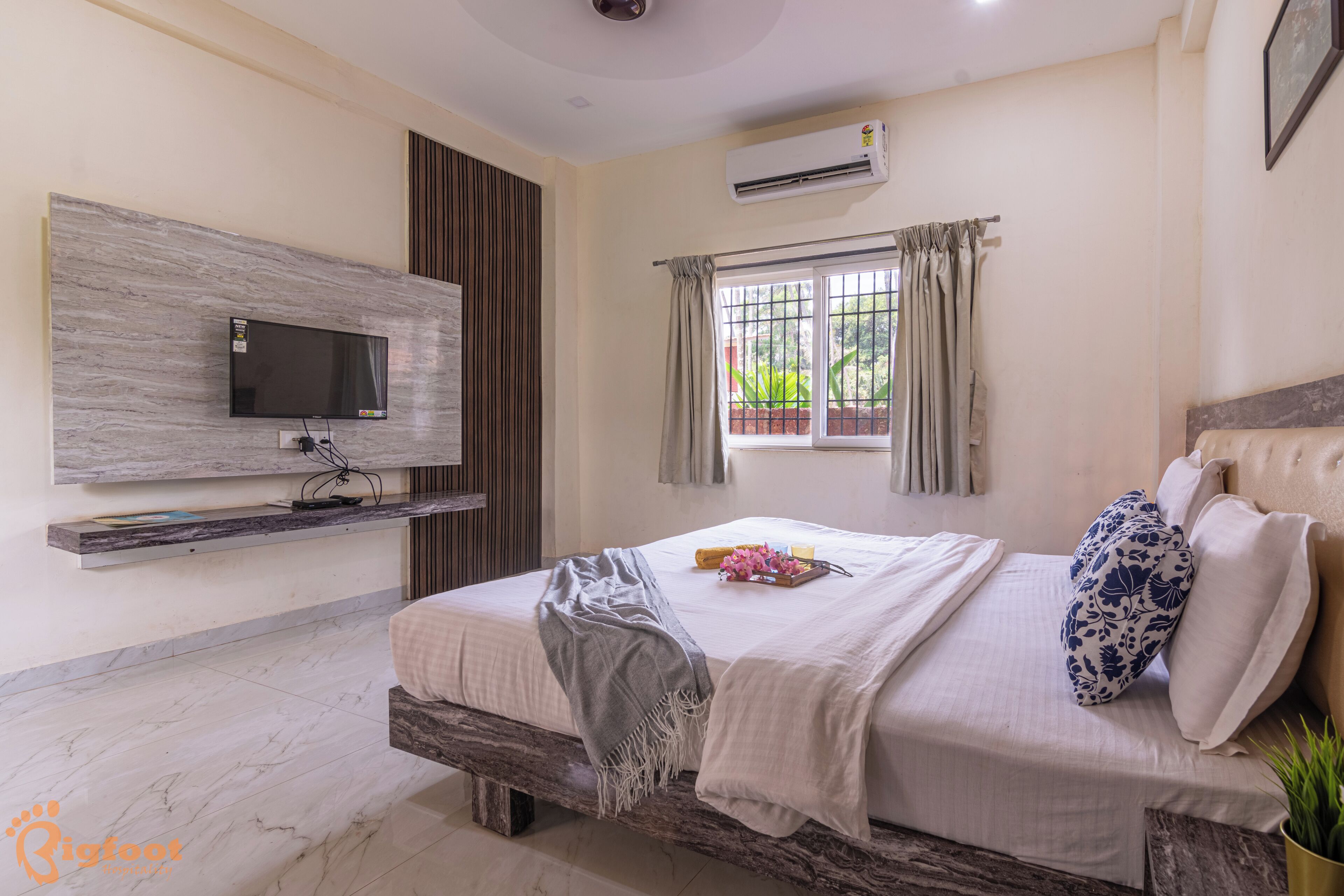 A bedroom with a bed, a mounted TV, a window with curtains, and an air conditioning unit.