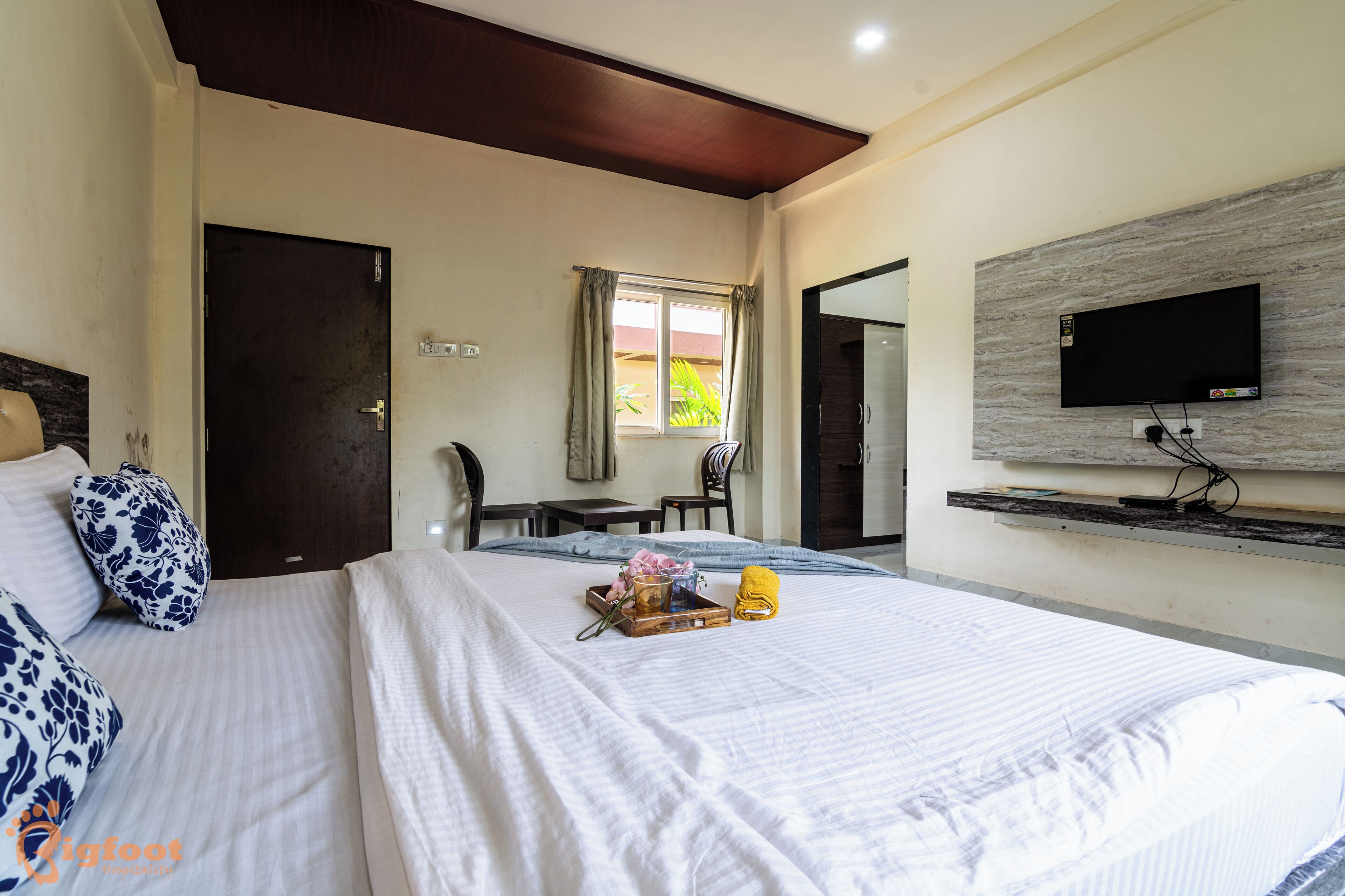 A hotel room with a bed, a TV mounted on the wall, a small table with chairs, and a window with curtains.