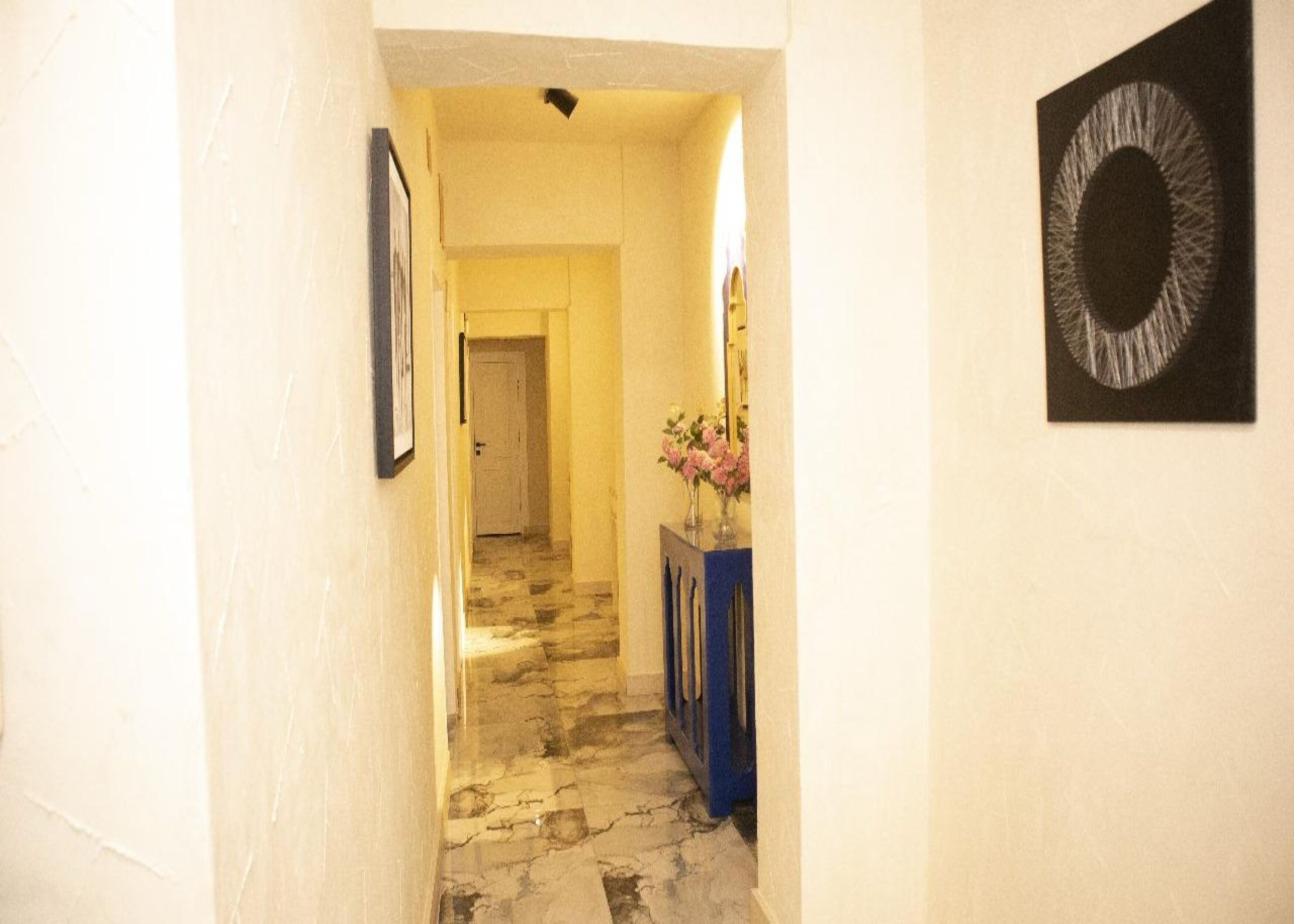 A hallway with a marble floor, a framed picture on the left, a vase with flowers on a blue console table, and a circular artwork on the right.