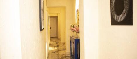 A hallway with a marble floor, a framed picture on the left, a vase with flowers on a blue console table, and a circular artwork on the right.