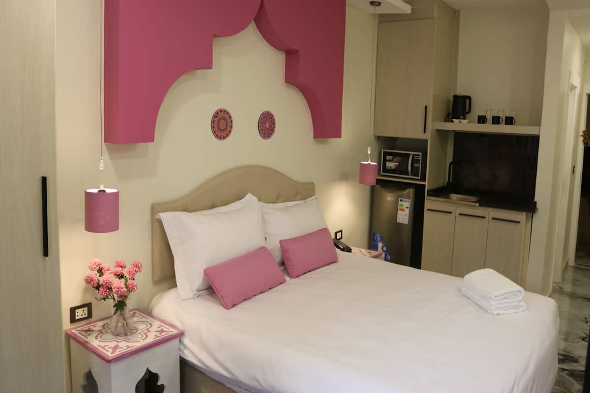 A hotel room with a bed, bedside table, lamp, and a kitchenette area.
