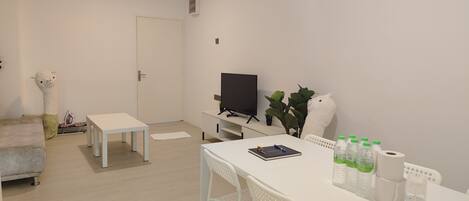 Una sala de estar moderna y minimalista con un sofá blanco, una mesita pequeña, un televisor sobre un mueble bajo y algunas plantas en macetas.