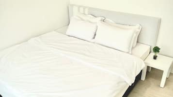 Una cama bien hecha con sábanas y almohadas blancas, una mesita de noche con una planta, y un suelo de madera clara.