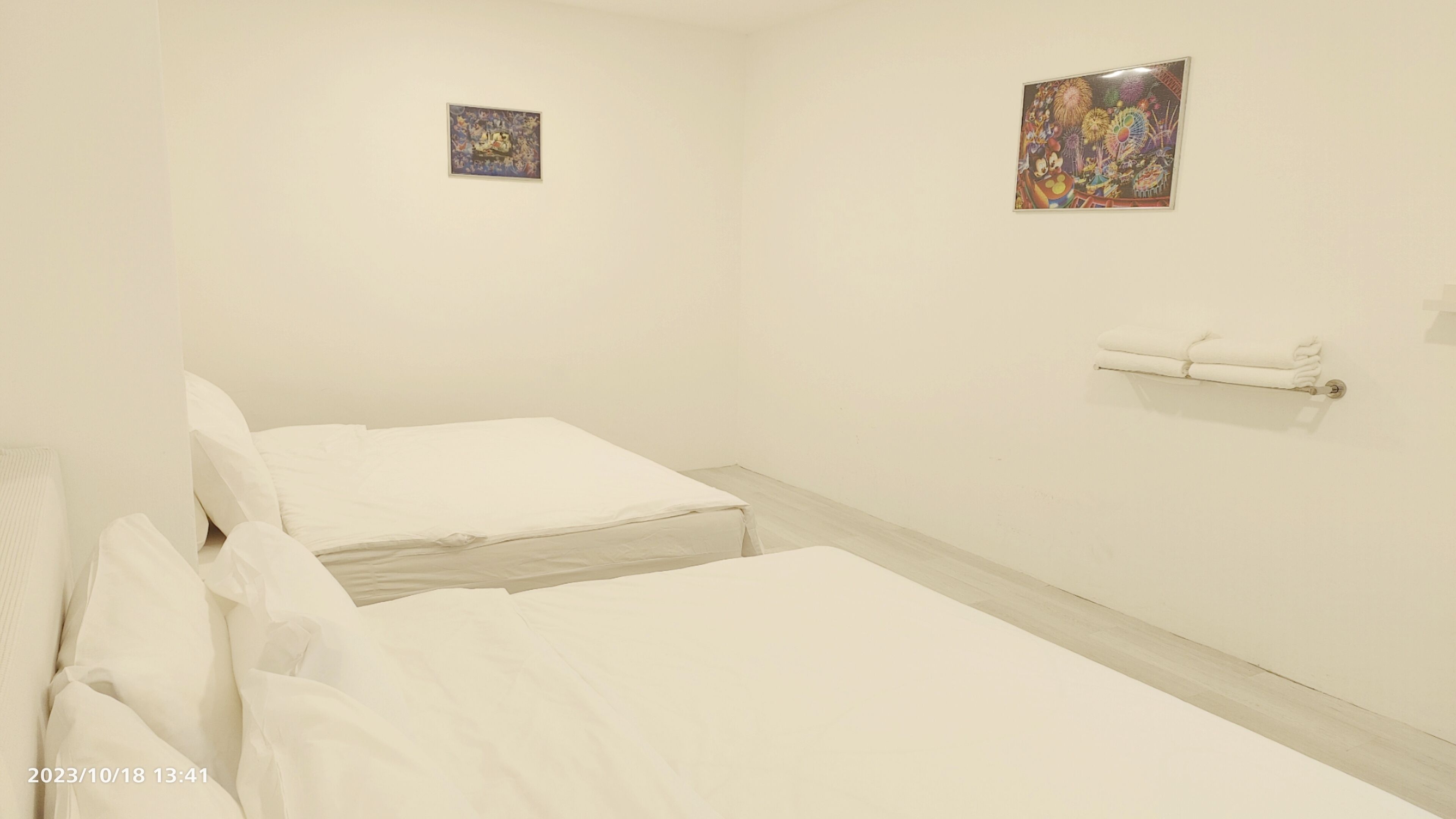 Ein schlicht eingerichtetes Hotelzimmer mit zwei Betten, einem kleinen Kunstwerk an der Wand und einem Handtuchhalter.
