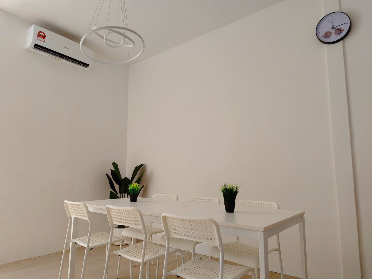 Un coin repas avec une table et des chaises blanches, un climatiseur fixé au mur et une horloge murale.