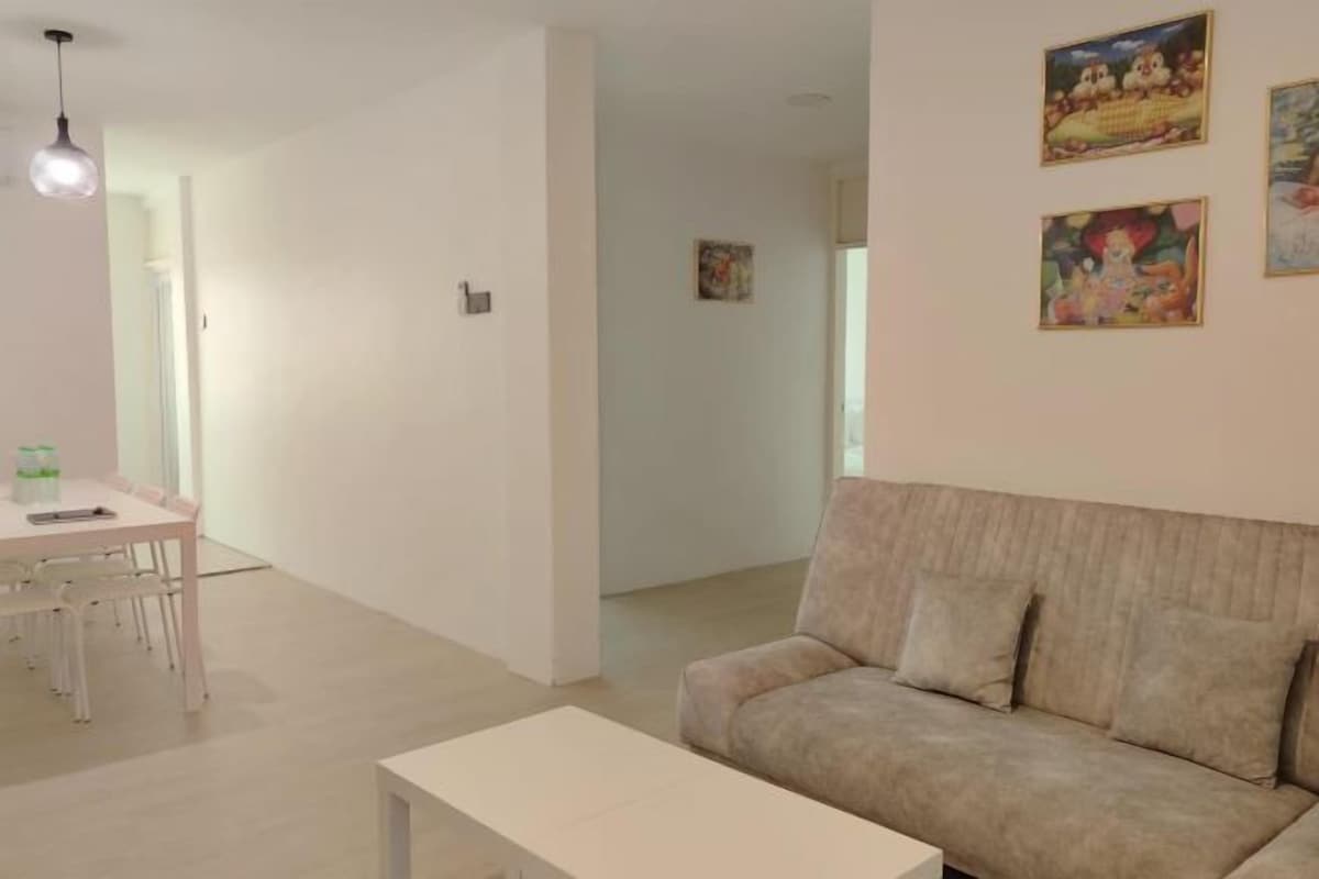 Un salon moderne avec un canapé beige, une table basse blanche et un coin repas comprenant une table et des chaises blanches. Deux tableaux sont accrochés au mur.