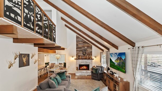 A spacious living room with a fireplace, a dining area, and a staircase leading to an upper level. - Revlands Cozy Cabin By Destination Big Bear: Close to Bear Mountain Ski Resort and Golf Course! Wood Burning Fireplace! Vaulted Ceilings! (Big Bear Lake)