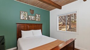 A bedroom with a wooden bed, a window with a view of trees, and three framed pictures on the wall. - Revlands Cozy Cabin By Destination Big Bear: Close to Bear Mountain Ski Resort and Golf Course! Wood Burning Fireplace! Vaulted Ceilings! (Big Bear Lake)