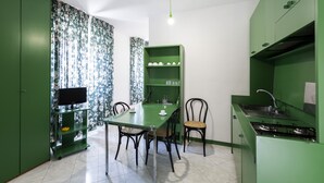 Eine kleine Küche mit Esstisch, Stühlen, einem grünen Schrank und einem Fernseher auf einem Standfuß.