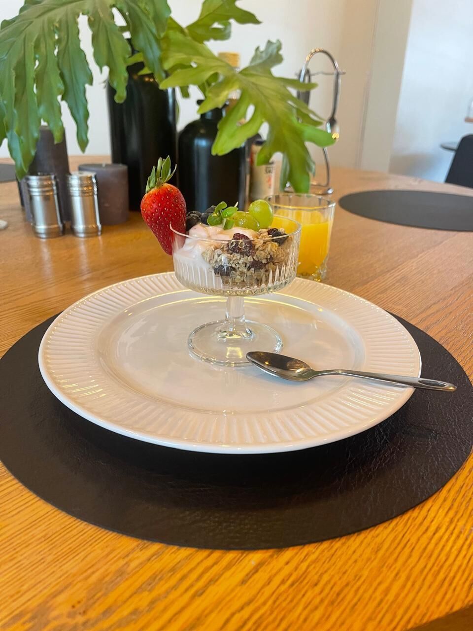 Ein Frühstückstisch mit einem Obstsalat in einer Glaskugel auf einem Teller, einem Löffel, einem Glas Orangensaft und einer Pflanze im Hintergrund.