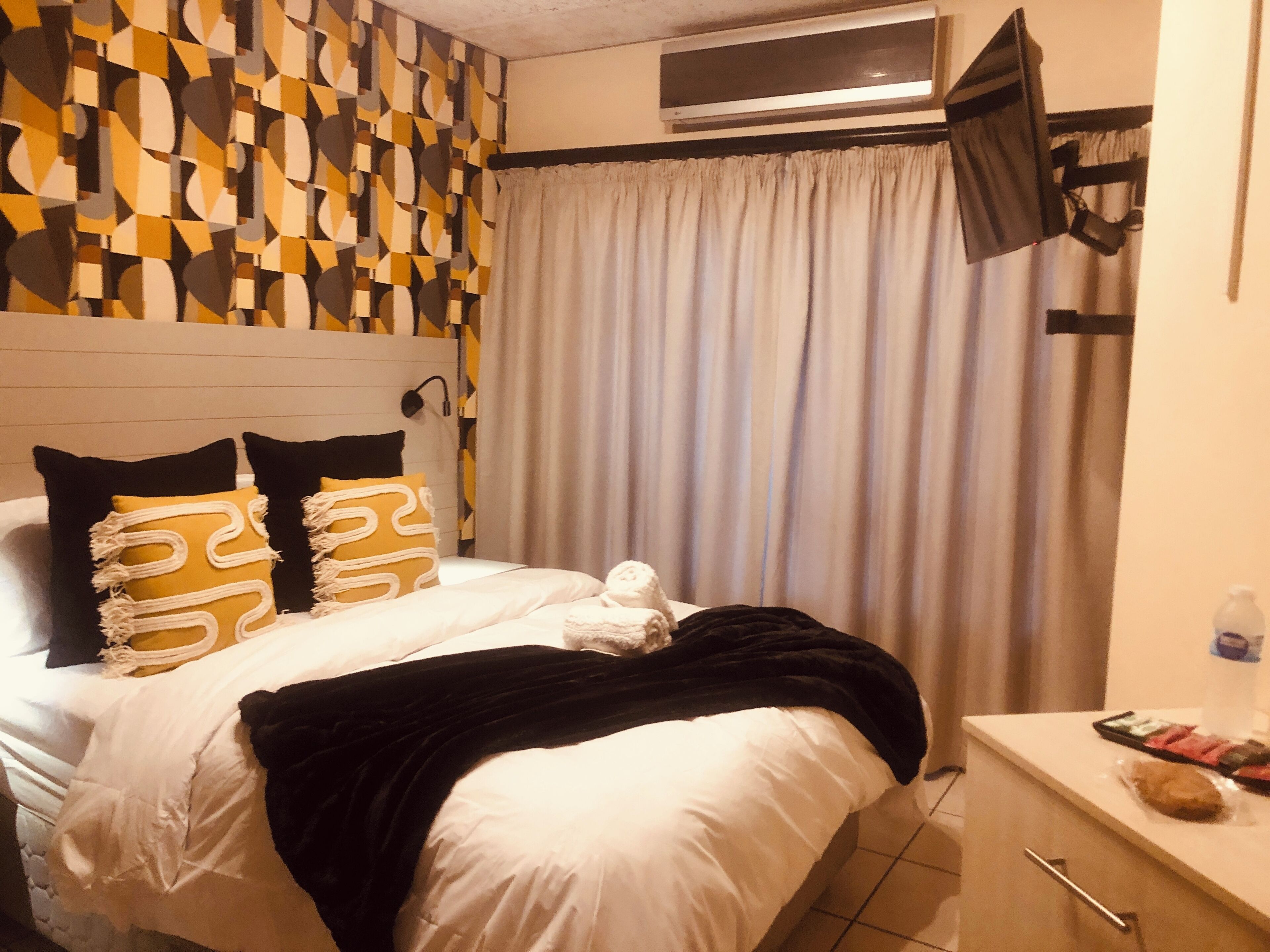 Une chambre d’hôtel avec un lit, un téléviseur à écran plat fixé au-dessus, un rideau et une petite table sur laquelle se trouvent une bouteille et une collation.