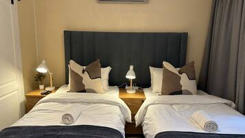 Habitación de hotel con dos camas, una mesita de noche con lámpara, aire acondicionado de pared y una ventana con cortinas.